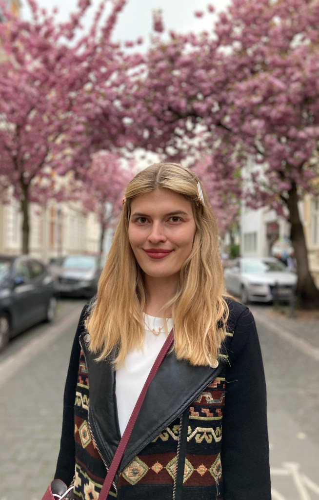 A photograph of a smiling person standing in a street with cherry blossom trees in the background.