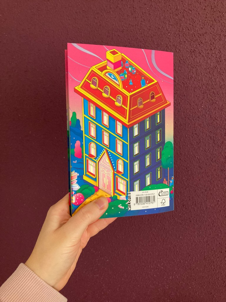 Hand holding up the book in front of a purple wall. The back of the book faces forward. On the back cover, there is a blue house with four stories, an attic and a chimney. The sky is pink and the style is cartoonish. 