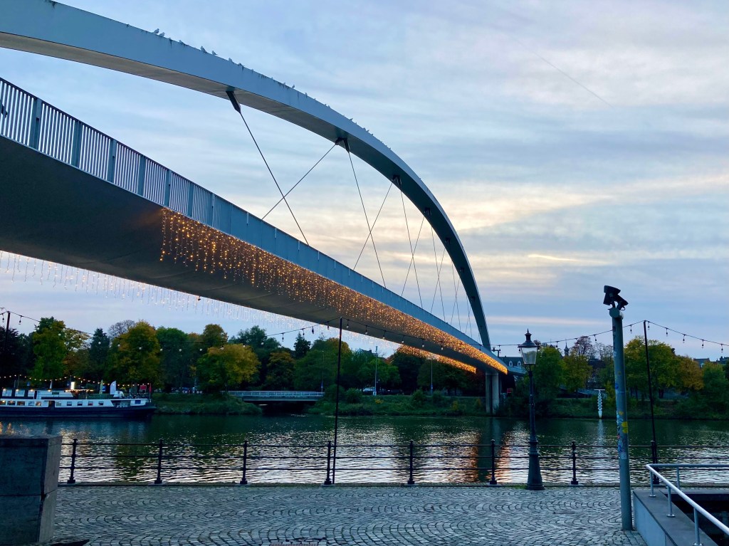A modern bridge spans a river. Golden fairy lights are handing from the sides of the bridge. The sky is cloudy but light. There is a lamppost on the embankment. A large boat is moored on the other side of the river.
